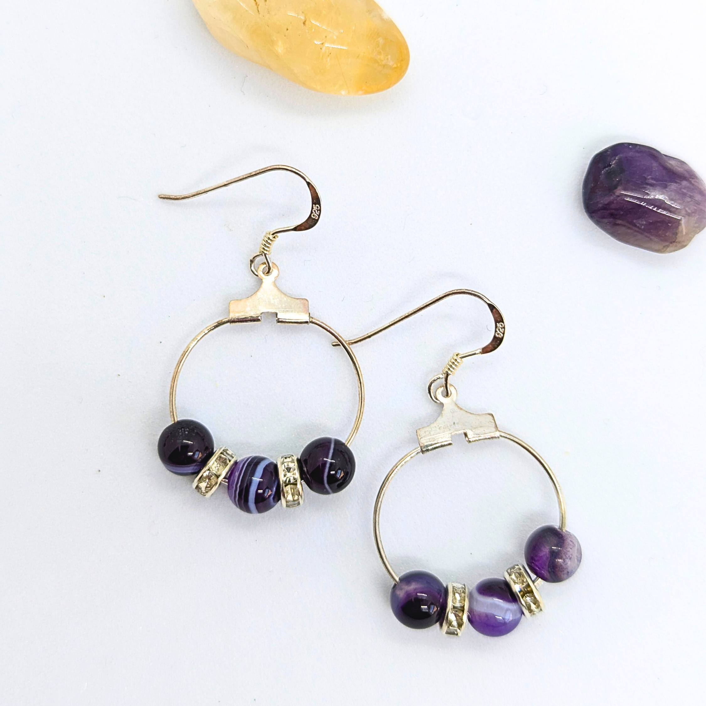 The Amethyst Statement Large Earrings by Silver Sunbird are a pair of circular, amethyst dangle earrings featuring three purple gemstones and two silver spacers each. The 925 sterling silver hooks complement the design, which is elegantly displayed on a white surface alongside a yellow crystal and a natural purple gemstone.