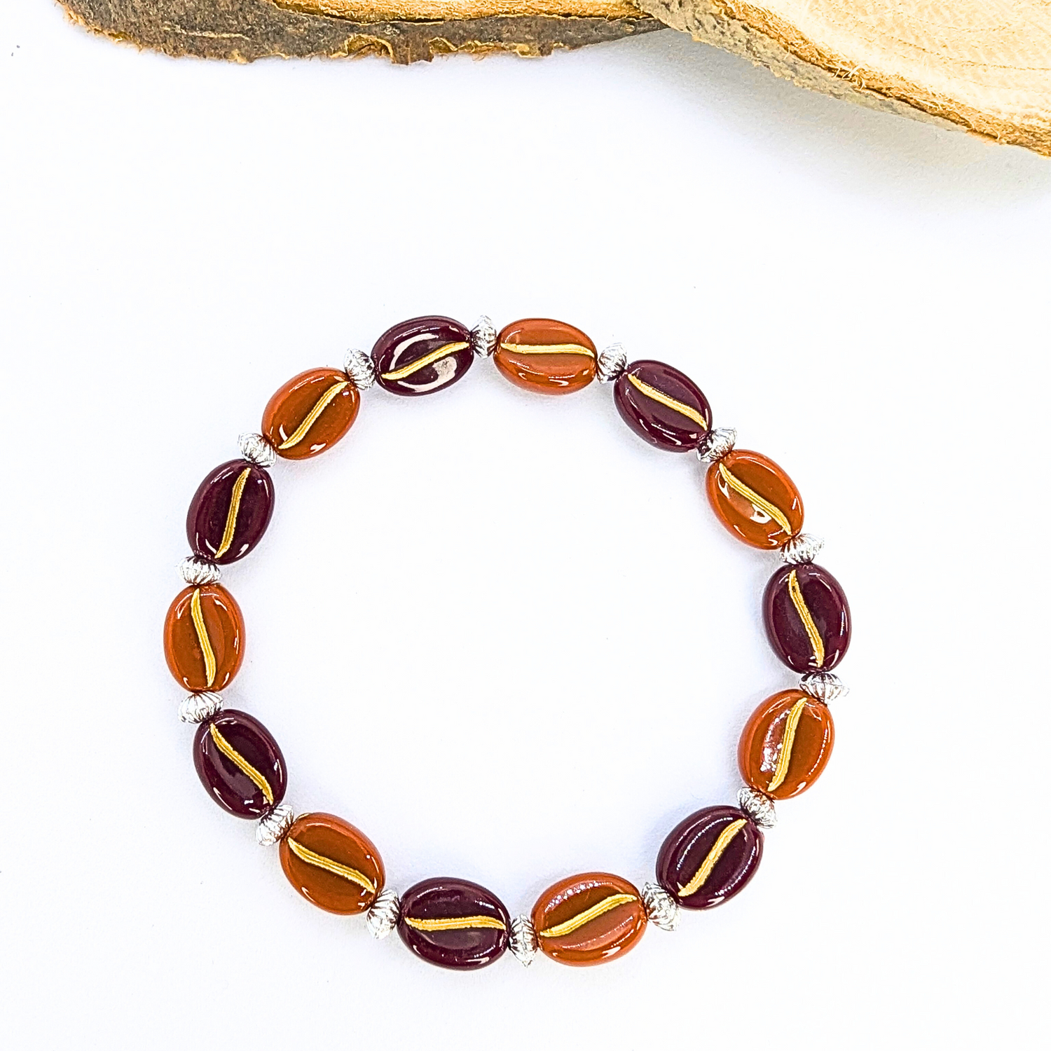 The Silver Sunbird Coffee Bean Bracelet elegantly combines red and orange oval beads with gold stripes, forming a charming circle reminiscent of coffee beans, all set against a subtle backdrop with hints of wood grain.