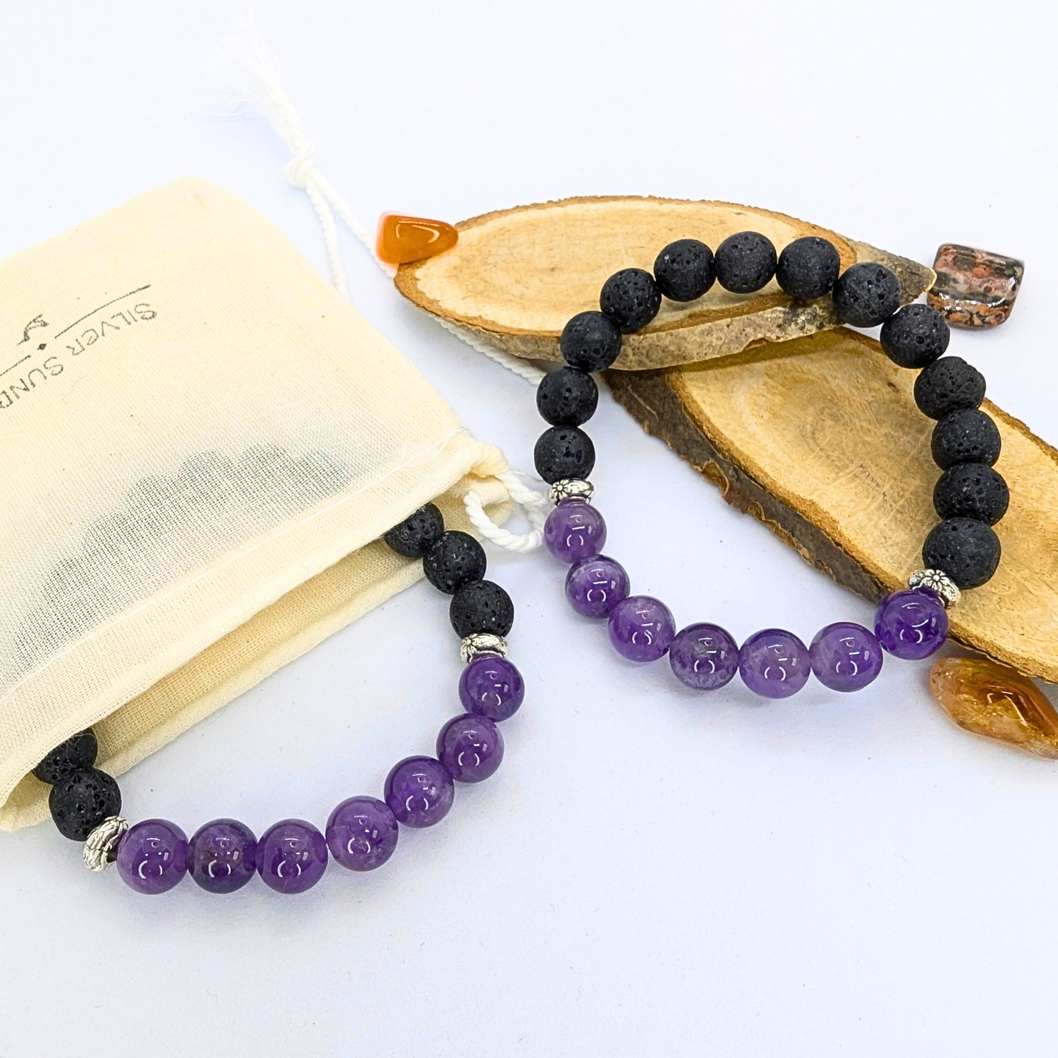 Two stretch bracelets featuring black lava stones and purple amethyst beads are displayed on a wooden slice. A beige drawstring pouch with &quot;Silver Surfer&quot; branding is positioned to the side, along with small polished stones in orange and brown hues. The amethyst beads are glossy and smooth, contrasting with the porous texture of the lava stones.