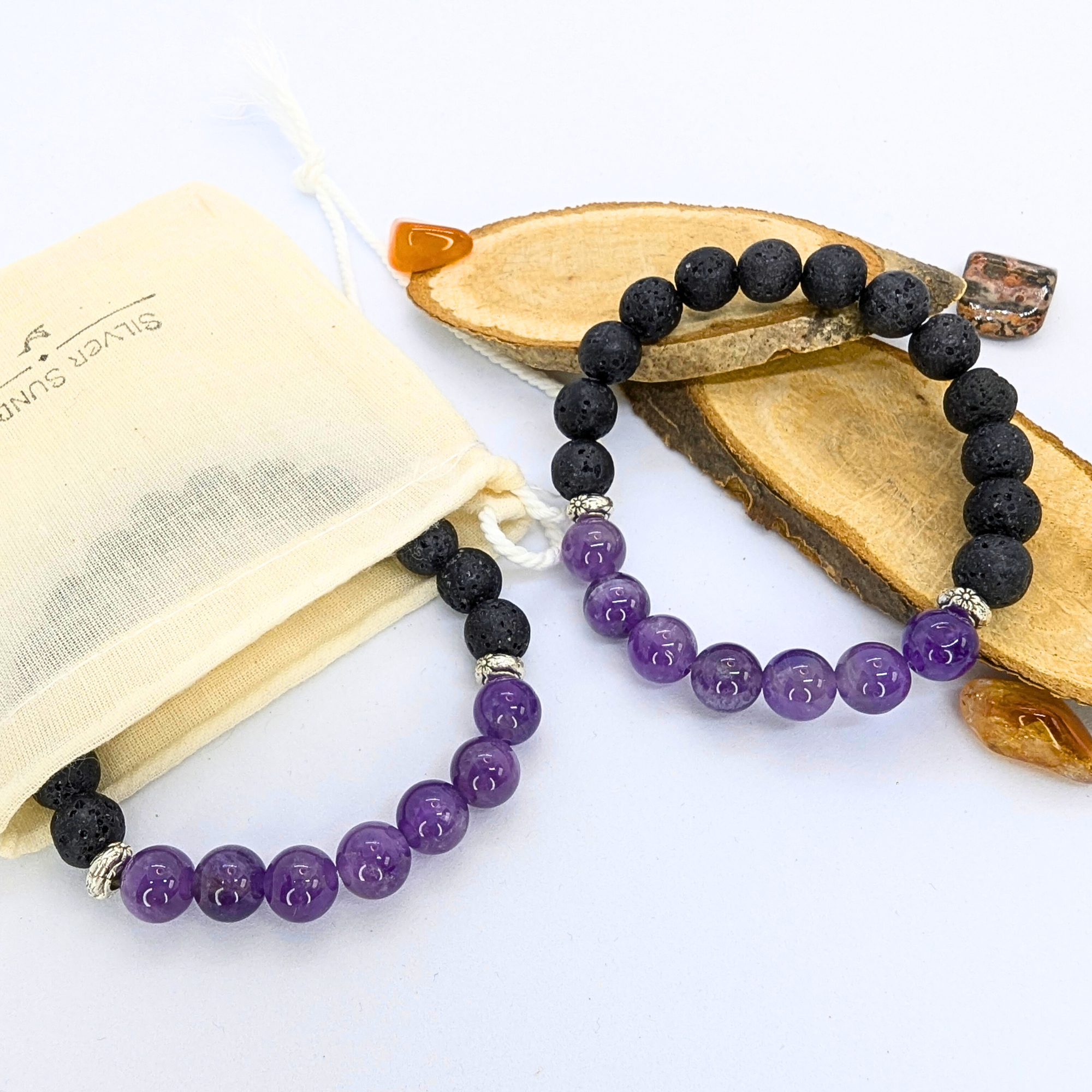 Two stretch bracelets featuring black lava stones and purple amethyst beads are displayed on a wooden slice. A beige drawstring pouch with &quot;Silver Surfer&quot; branding is positioned to the side, along with small polished stones in orange and brown hues. The amethyst beads are glossy and smooth, contrasting with the porous texture of the lava stones.