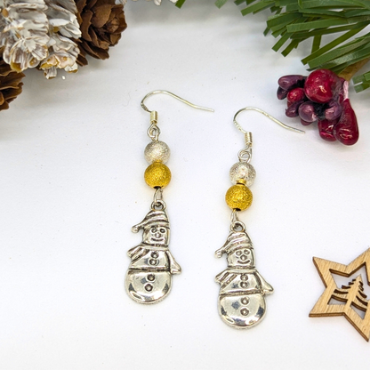 Very Merry Snowmen Earrings by Silver Sunbird: Handmade earrings with silver snowman charms and gold &amp; silver sparkle beads, shown on a white surface with pine cones, greenery, and a wooden star ornament.