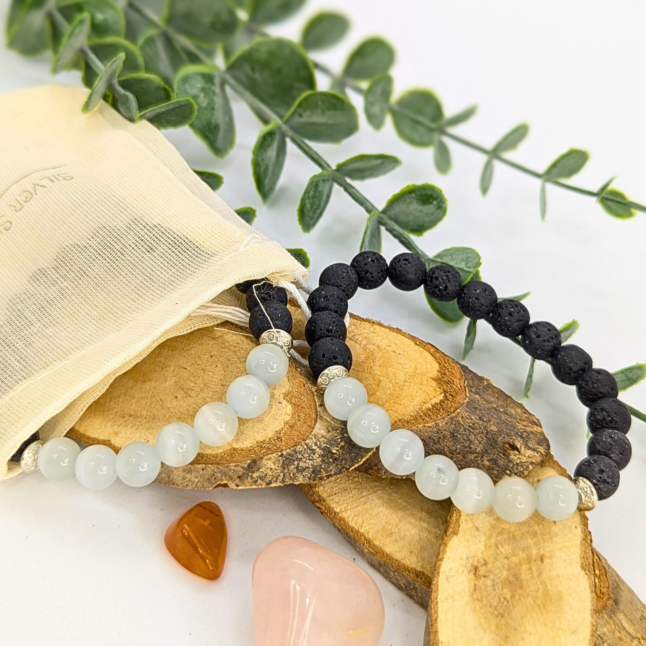 The Moonstone and Lava Bracelets by Silver Sunbird rest on wooden pieces next to a cream pouch, surrounded by green leaves, an orange stone, and a pink crystal for a peaceful, natural display.