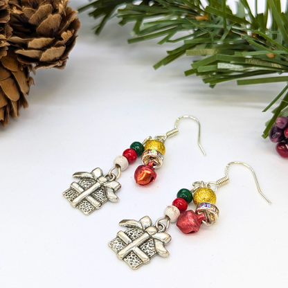 The Silver Sunbird Gift Box Dangle Earrings feature silver present charms, red and green beads, gold accents, and sterling silver hooks, shown on a white background with pinecones and evergreen branches.