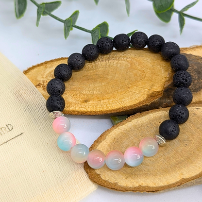The Silver Sunbird Mermaid-Inspired Lava Stone Bracelet features alternating black lava beads and pastel pink, blue, and white stones—perfect for ocean lovers. Shown on wooden slices with green leaves in the background.