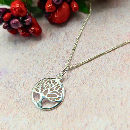 The Silver Sunbird Tree of Life Sterling Silver Necklace features a round pendant, displayed on a light textured surface with red and purple artificial berries—an elegant, meaningful accessory for any occasion.
