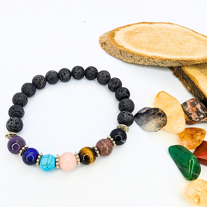 The Chakra Bracelet by Silver Sunbird, featuring black lava stones and colorful gemstones, is shown beside polished healing stones and two cut wood pieces on a white background.