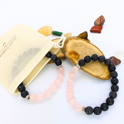 A Rose Quartz and Lava Stone Bracelet from Silver Sunbird rests on a wooden slice, accompanied by an assortment of small crystals. Nearby, an open fabric pouch reveals another similar bracelet, all set against a white background.