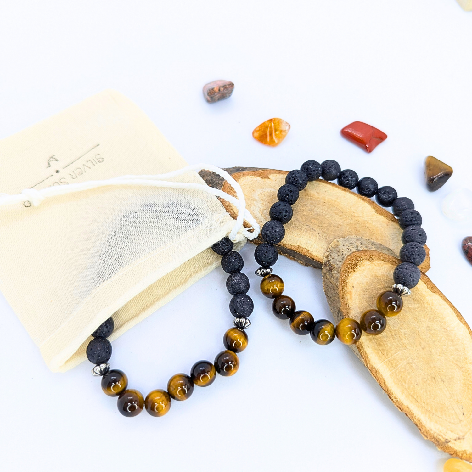 The Tiger Eye and Lava Stone Bracelets by Silver Sunbird are elegantly showcased on wooden pieces with a beige drawstring pouch. The display features two beaded bracelets of black and brown beads, set against a white background with colorful stones scattered around.