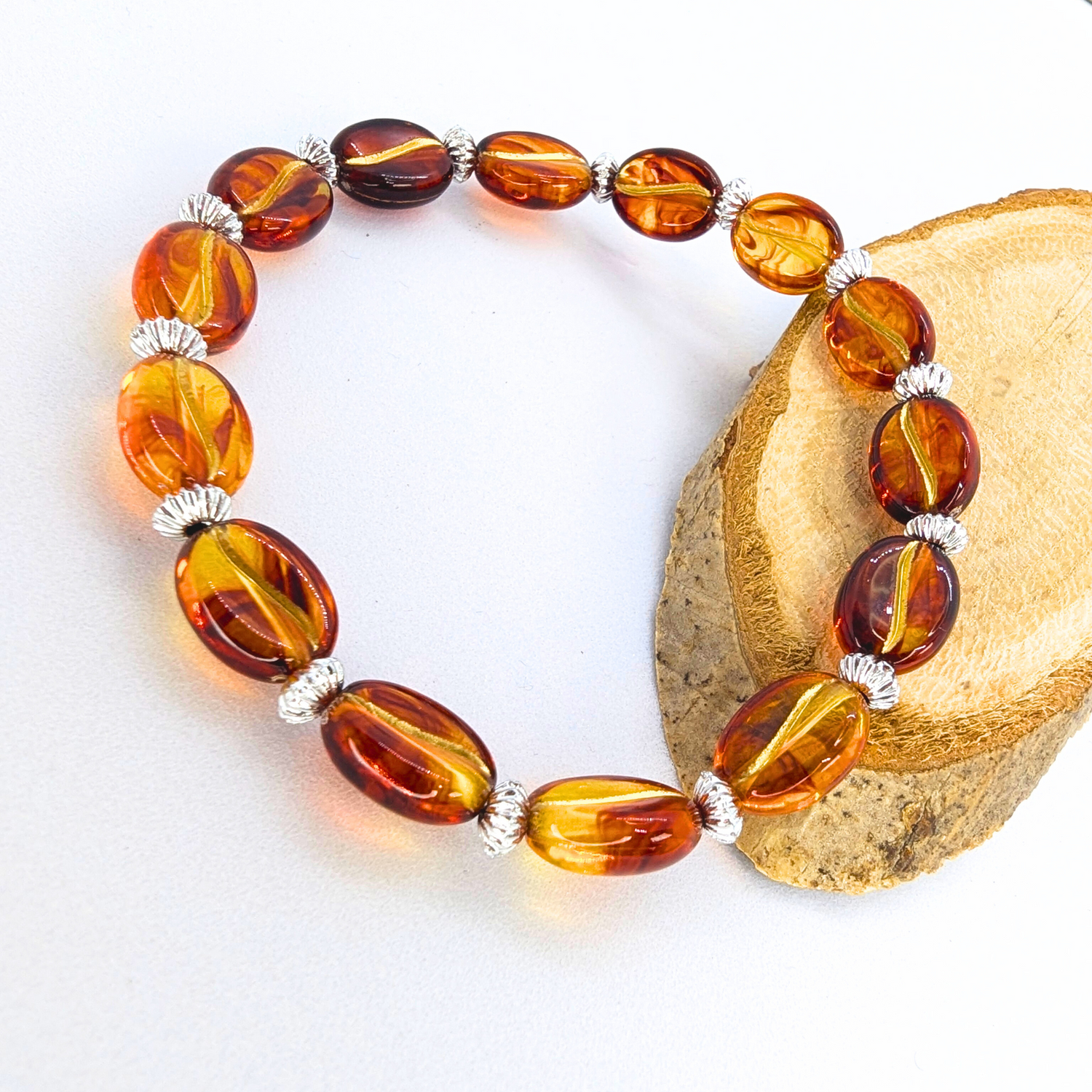 The Silver Sunbird Coffee Bean Bracelet features oval amber beads in orange and brown hues with small silver spacers, elegantly arranged in a circle and catching the light beautifully on a light wooden surface.