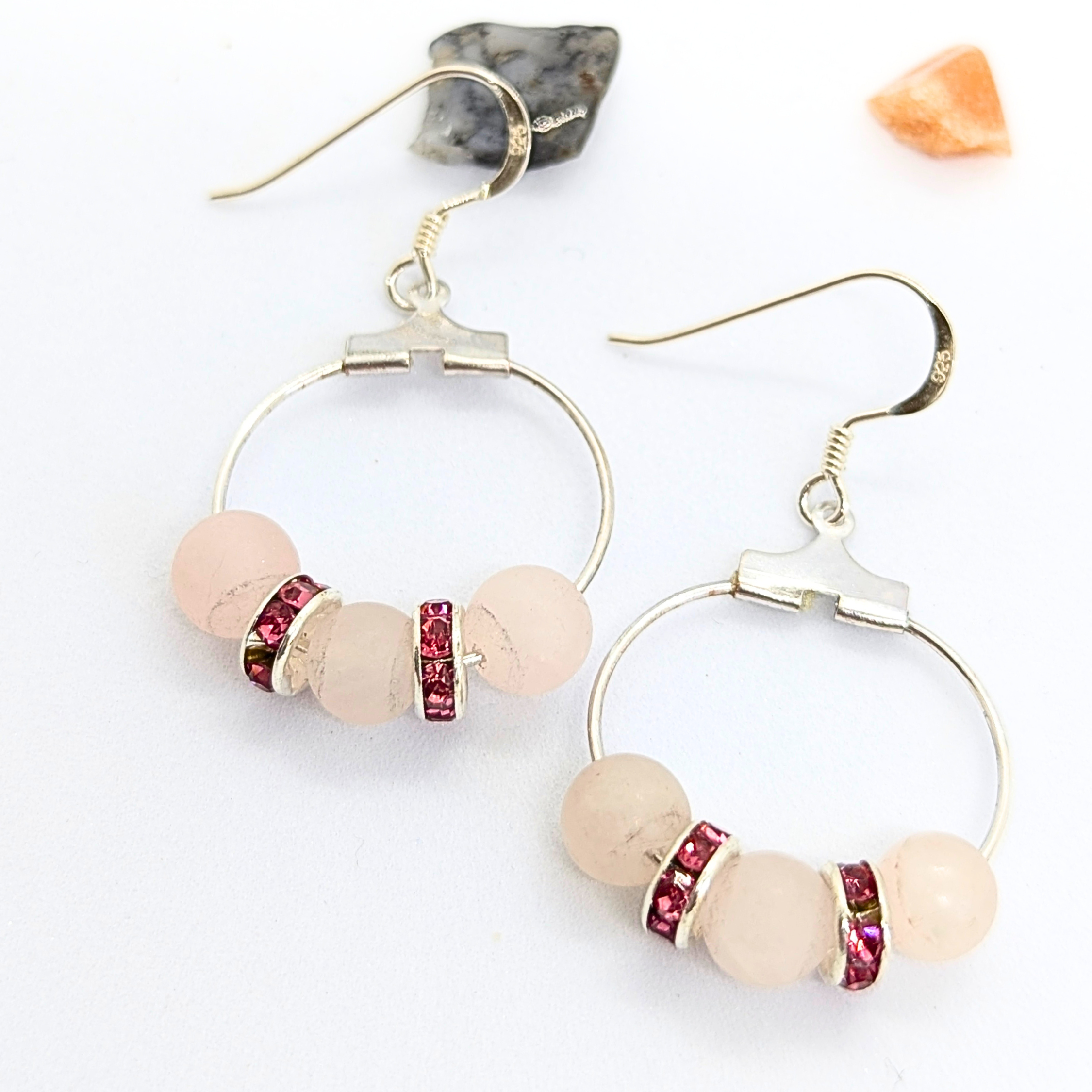 Introducing the Rose Quartz Statement Earrings by Silver Sunbird: These hypoallergenic sterling silver hoop earrings feature round rose quartz beads, elegantly separated by pink rhinestone spacers. Laid out with a piece of raw gemstone and an orange crystal in the background, the earrings are completed with elegant silver hooks that showcase an exquisite design.