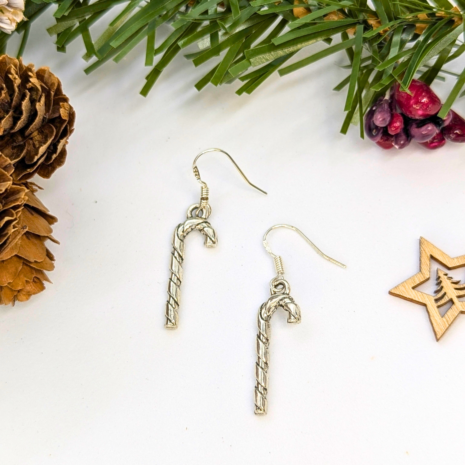 Classic Candy Cane Earrings by Silver Sunbird are displayed on a white surface, accented with pine needles, a pine cone, red berries, and a wooden star ornament for a festive Christmas jewelry setting.