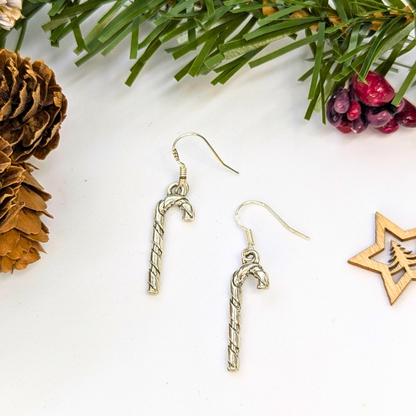 Classic Candy Cane Earrings by Silver Sunbird are displayed on a white surface, accented with pine needles, a pine cone, red berries, and a wooden star ornament for a festive Christmas jewelry setting.