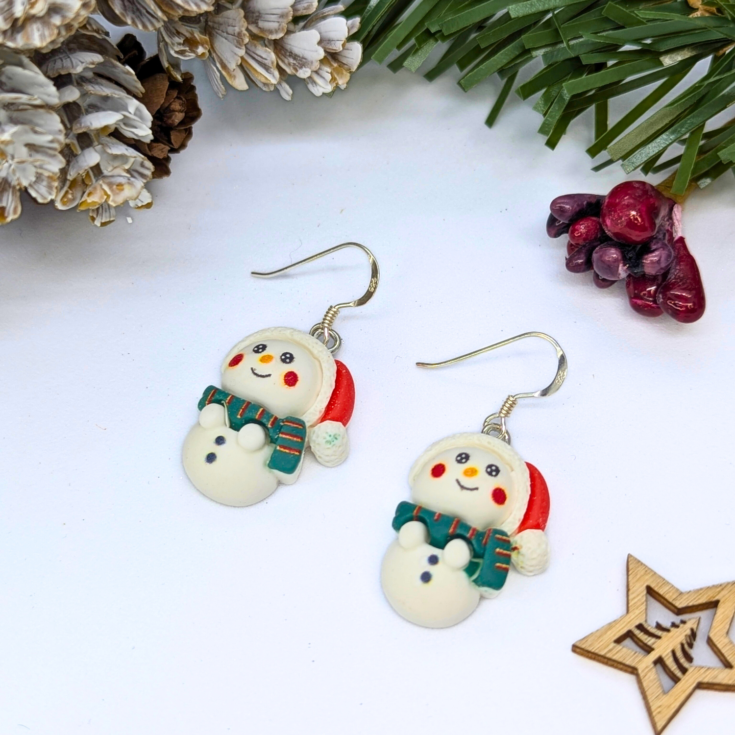 Bright Snowmen Earrings by Silver Sunbird feature red hats and green scarves, displayed on a white surface with pine branches, frosted pinecones, berries, and a wooden star—ideal Christmas jewelry for holiday enthusiasts.