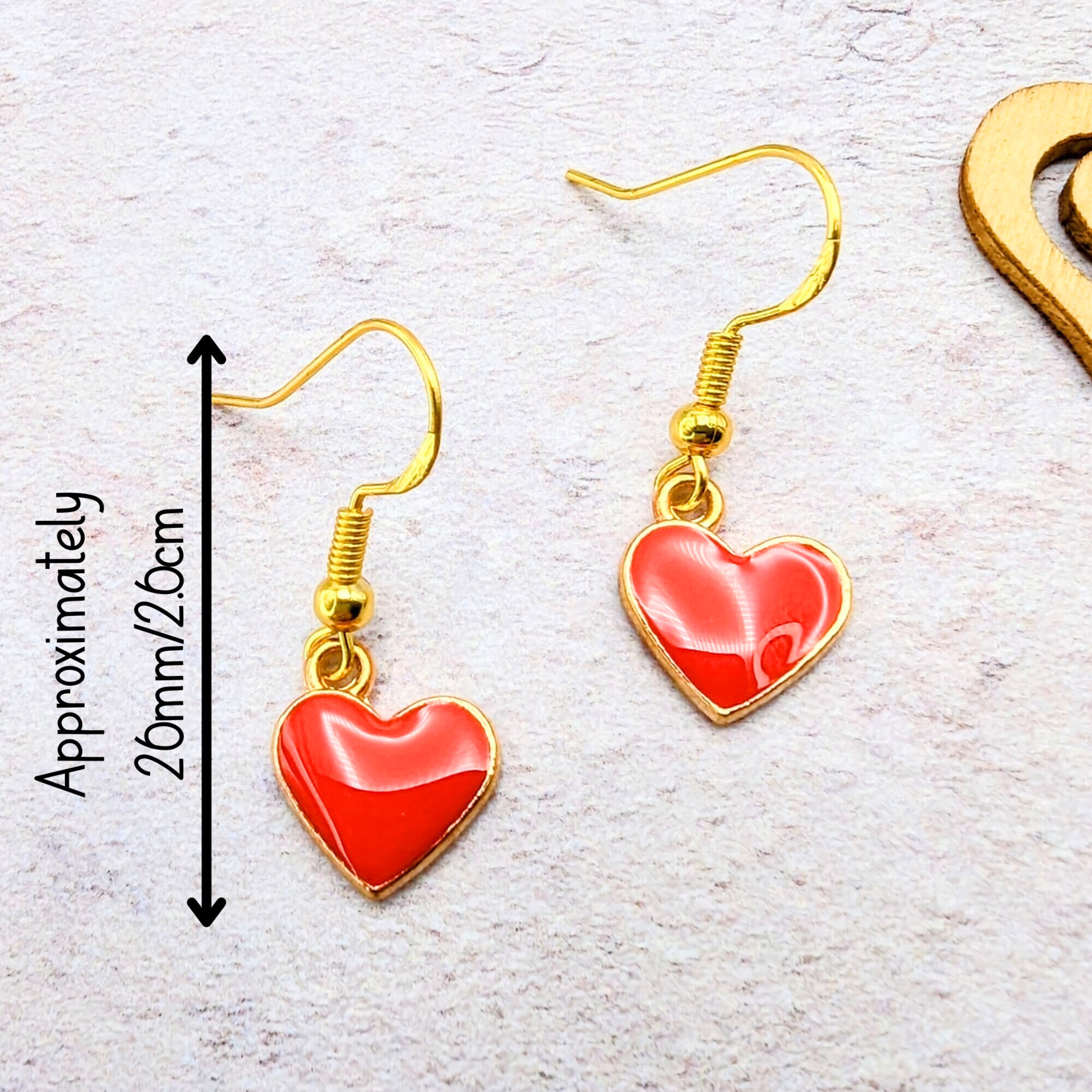 The Alluring Red Heart Earrings by Silver Sunbird feature gold hooks and red heart-shaped pendants, perfect as hypoallergenic earrings. Each measures approximately 20mm (2cm). They are beautifully displayed on a textured light surface.