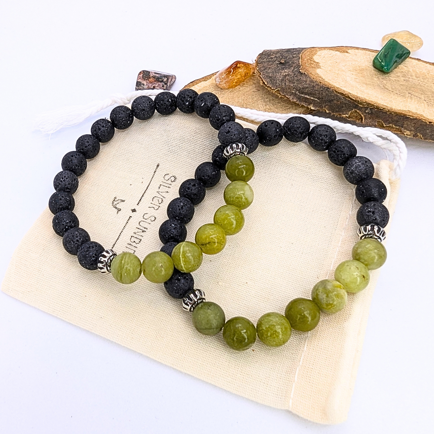 Two beaded bracelets from Silver Sunbird rest on a wooden slice: one black with silver accents and the other a green Peridot Bracelet. They are in a cloth pouch, surrounded by small gemstones, showcasing the charm of Silver Sunbird&