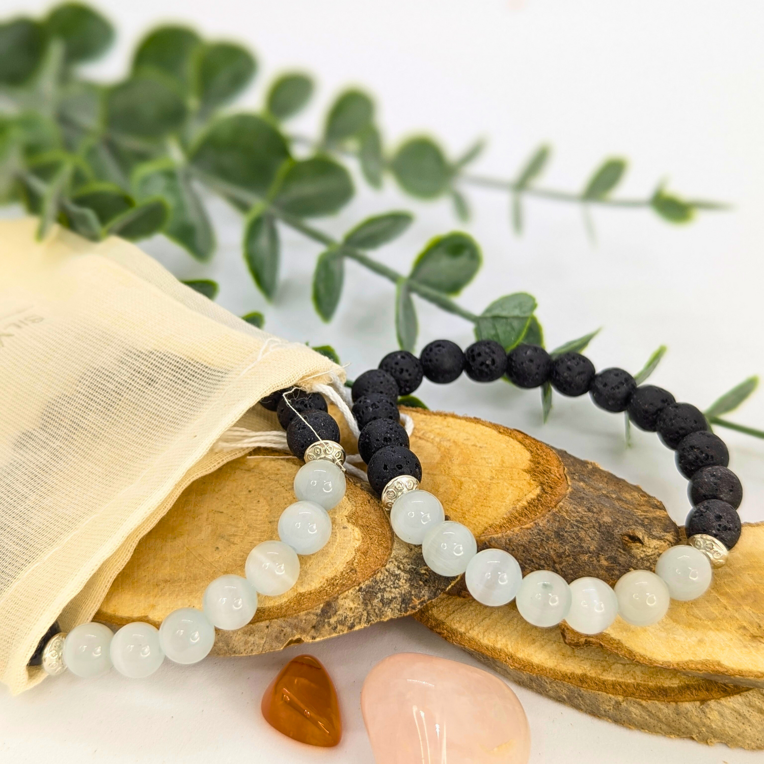The Silver Sunbird Moonstone and Lava Bracelets—one featuring white moonstone beads, the other black lava stone—are displayed on wood slices with greenery, accompanied by a cream pouch and two polished stones.