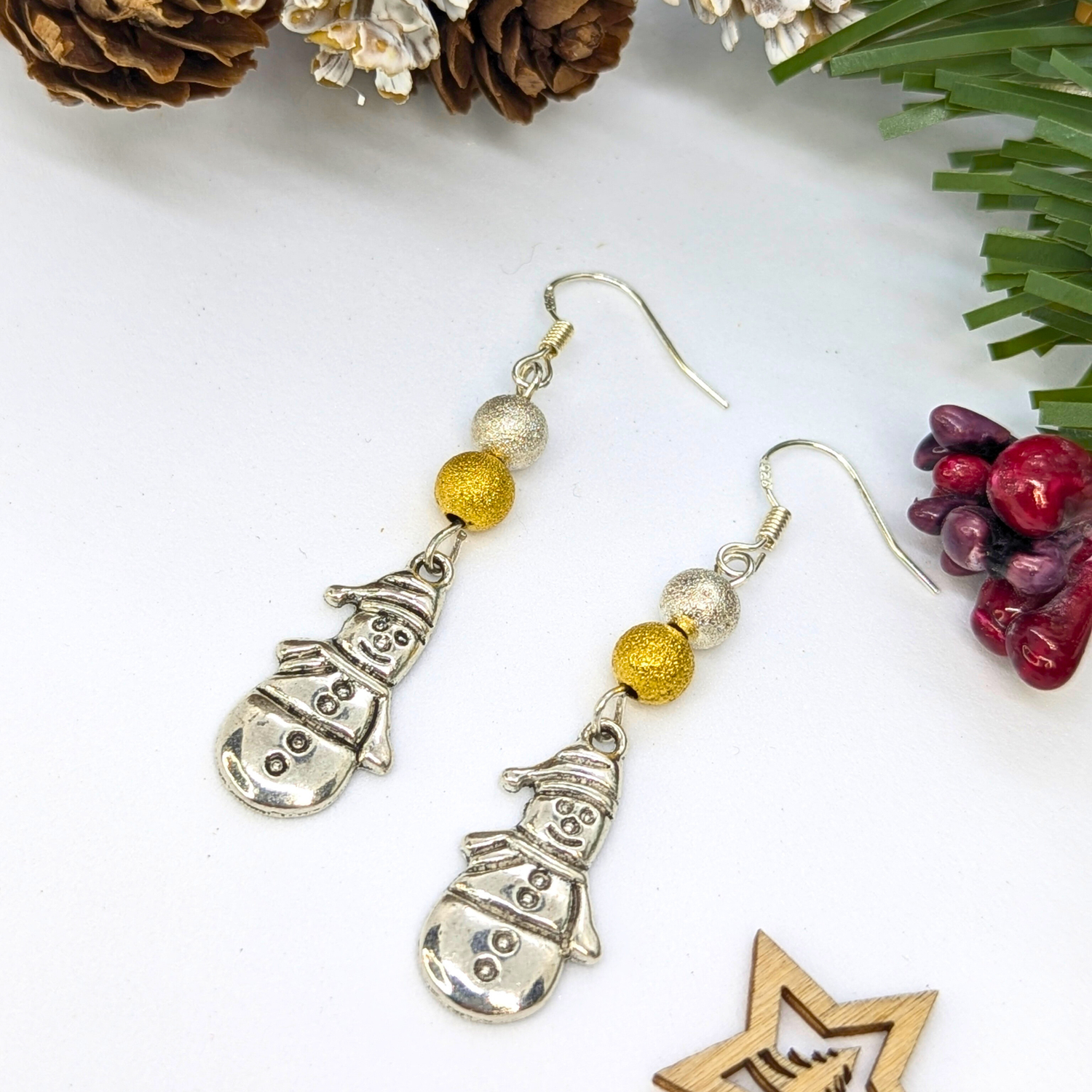 The Silver Sunbird Very Merry Snowmen Earrings feature handmade silver snowman charms with gold and silver glitter beads, displayed festively with pinecones, faux pine branches, and red berries on a white surface.