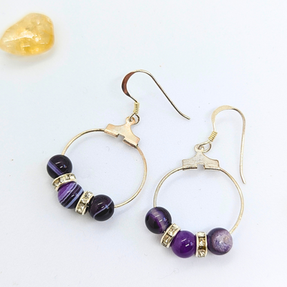 The Silver Sunbird Amethyst Statement Large Earrings, featuring gold-tone hooks and circular frames, are adorned with three purple amethyst and two silver rhinestone spacers. A small yellow natural gemstone is positioned near the top left corner of the white background.