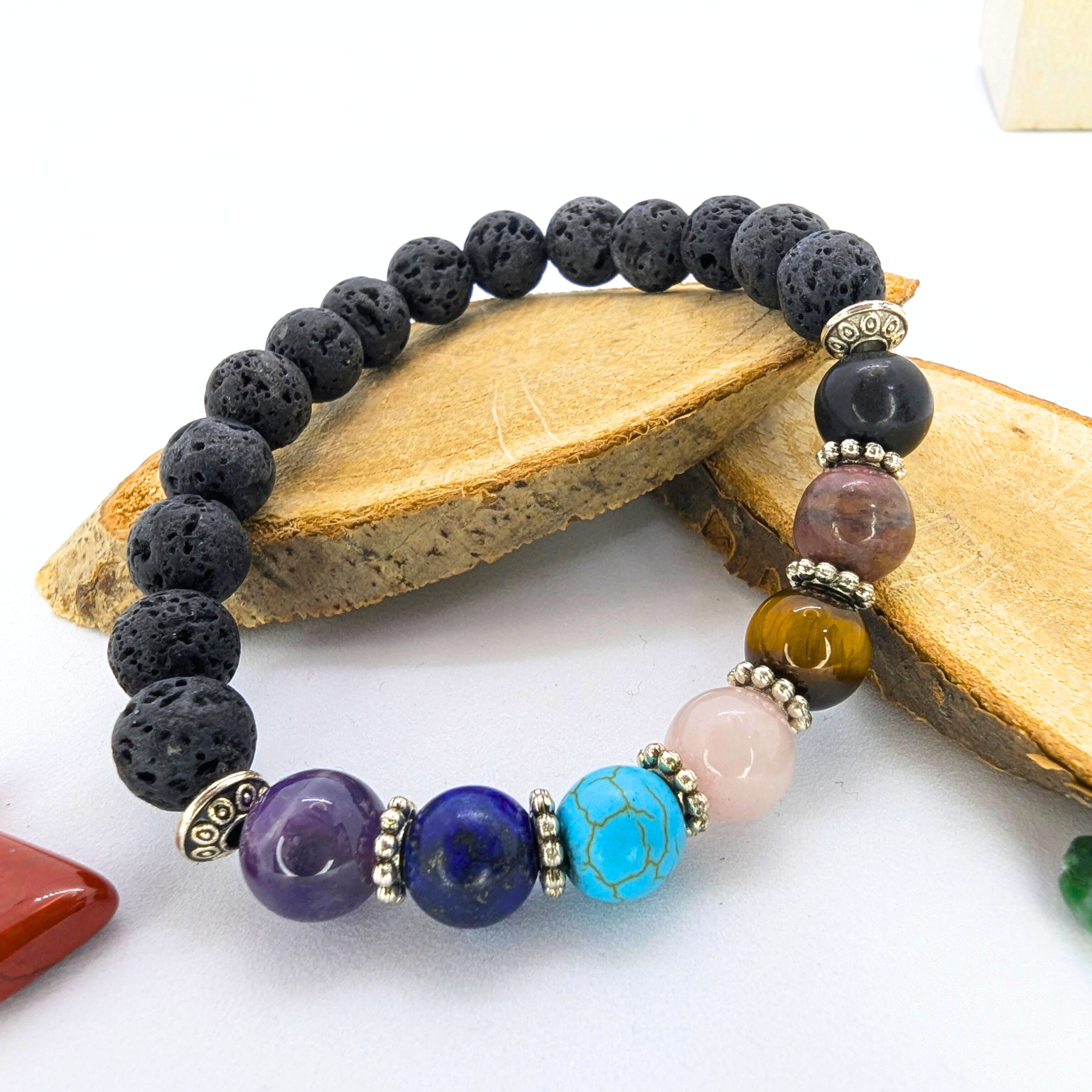The Chakra Bracelet by Silver Sunbird features black lava stones, multicolored semi-precious gems, and silver spacers. Displayed on two wooden pieces against a white background, with polished stones scattered nearby.