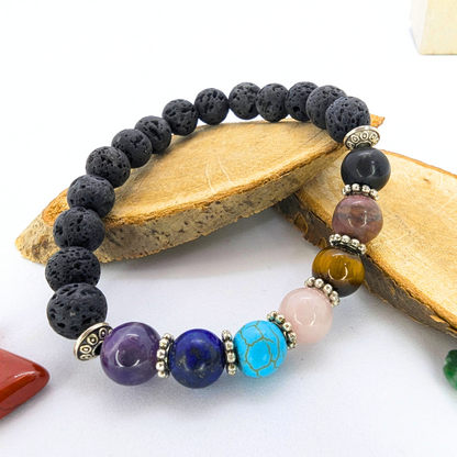 The Chakra Bracelet by Silver Sunbird features black lava stones, multicolored semi-precious gems, and silver spacers. Displayed on two wooden pieces against a white background, with polished stones scattered nearby.
