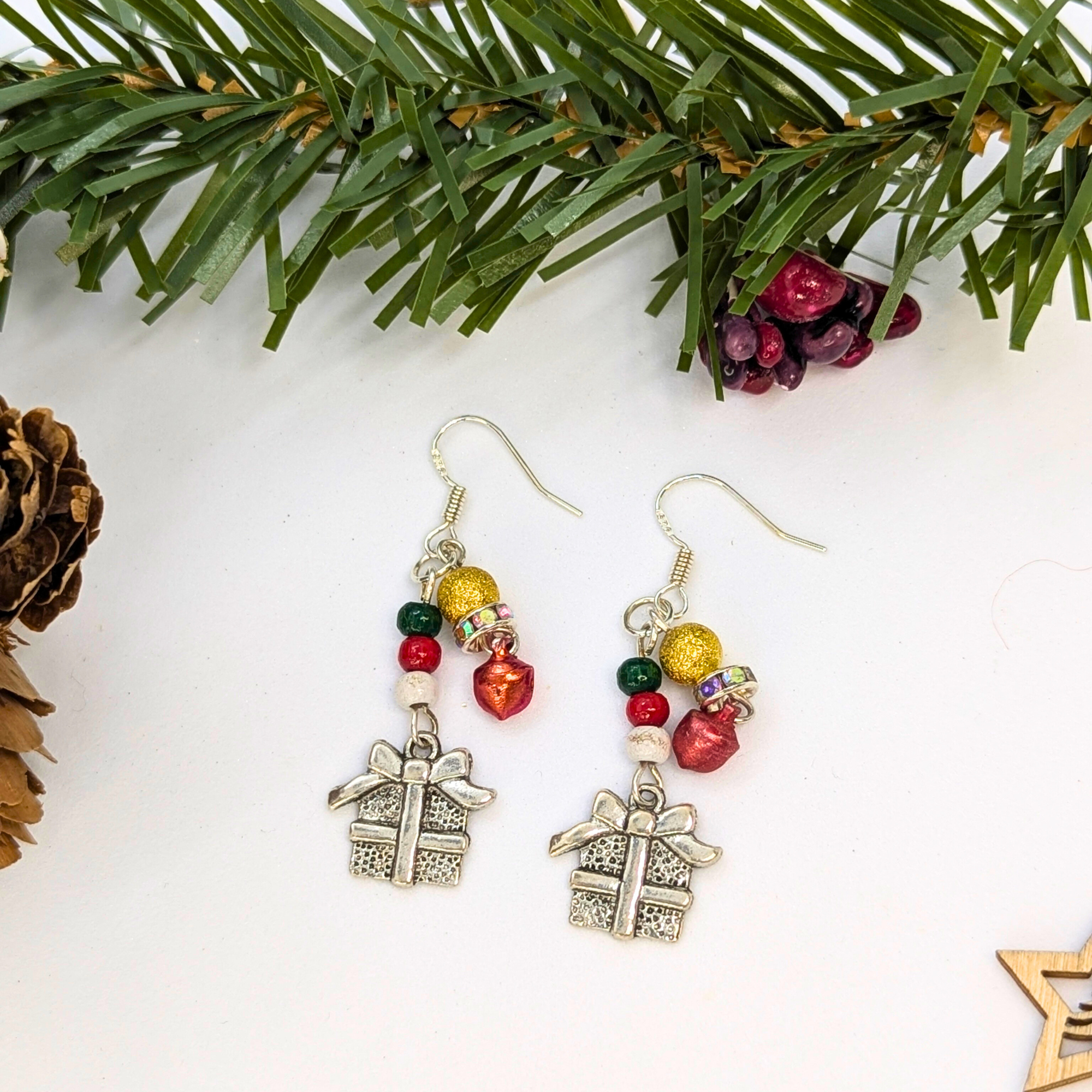 The Silver Sunbird Gift Box Dangle Earrings feature sterling silver hooks and red, green, and gold beads. Displayed with festive pine branches, they make the perfect Christmas accessory!.