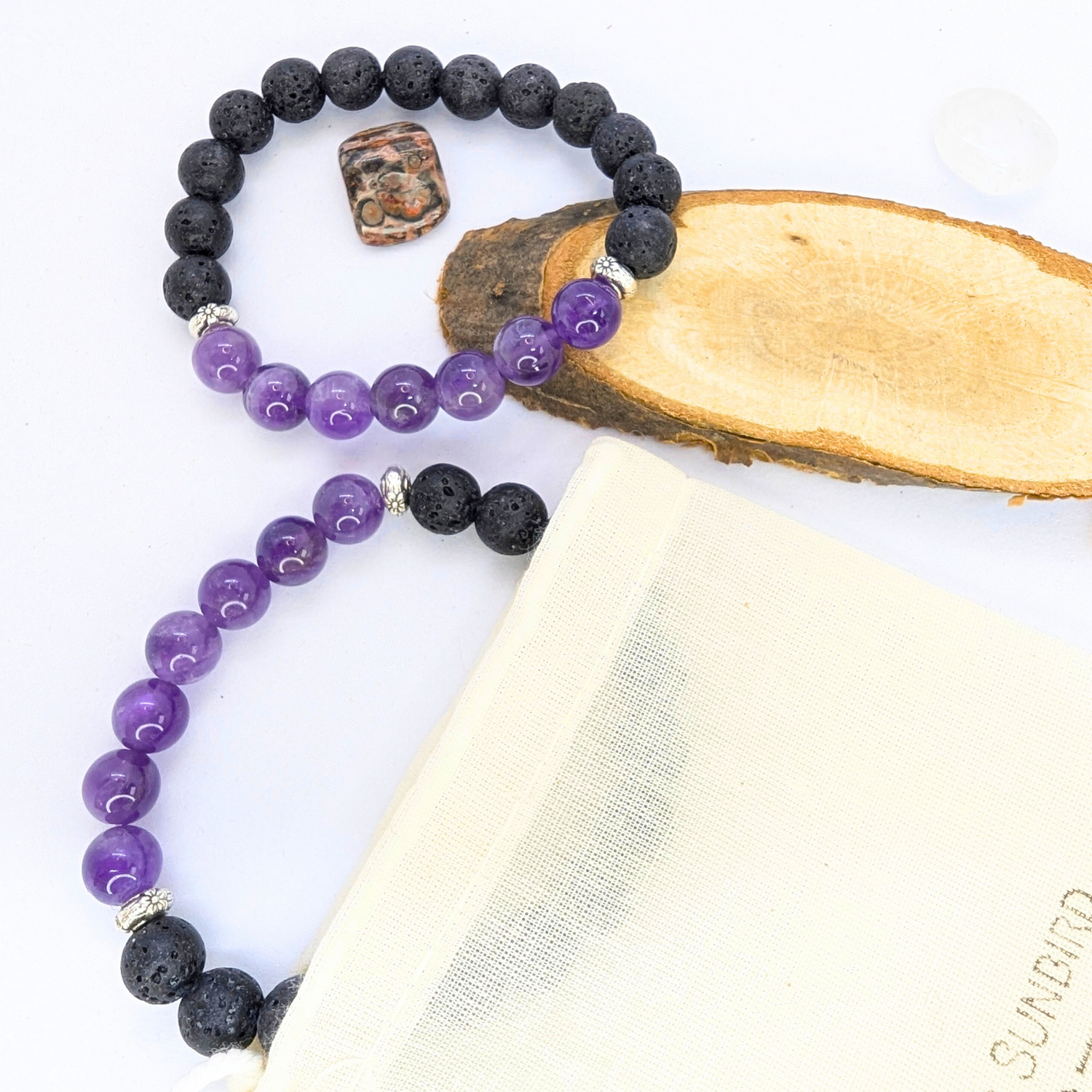 Two stretch bracelets featuring black lava stones and purple amethyst beads are displayed on a wooden slice. A beige drawstring pouch with &quot;Silver Surfer&quot; branding is positioned to the side, along with small polished stones in orange and brown hues. The amethyst beads are glossy and smooth, contrasting with the porous texture of the lava stones.