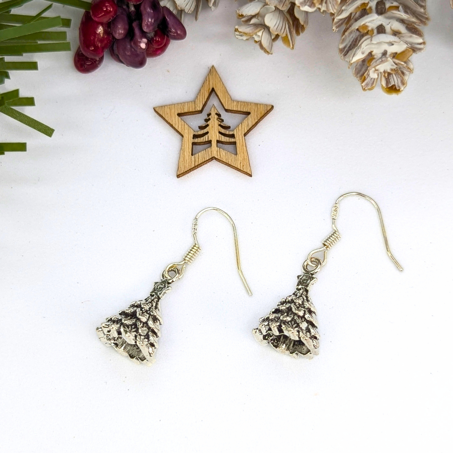 A pair of Silver Sunbird 3D Christmas Tree Earrings rests on a white surface beside a wooden star ornament with a tree cutout, surrounded by pine cones, berries, and greenery for a festive holiday atmosphere.
