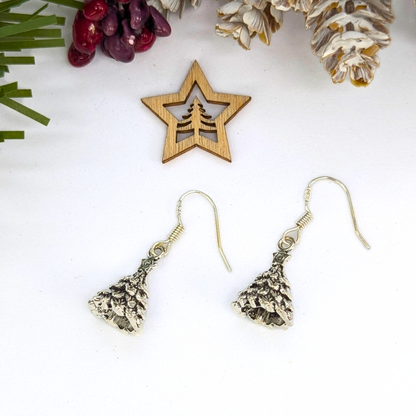 A pair of Silver Sunbird 3D Christmas Tree Earrings rests on a white surface beside a wooden star ornament with a tree cutout, surrounded by pine cones, berries, and greenery for a festive holiday atmosphere.