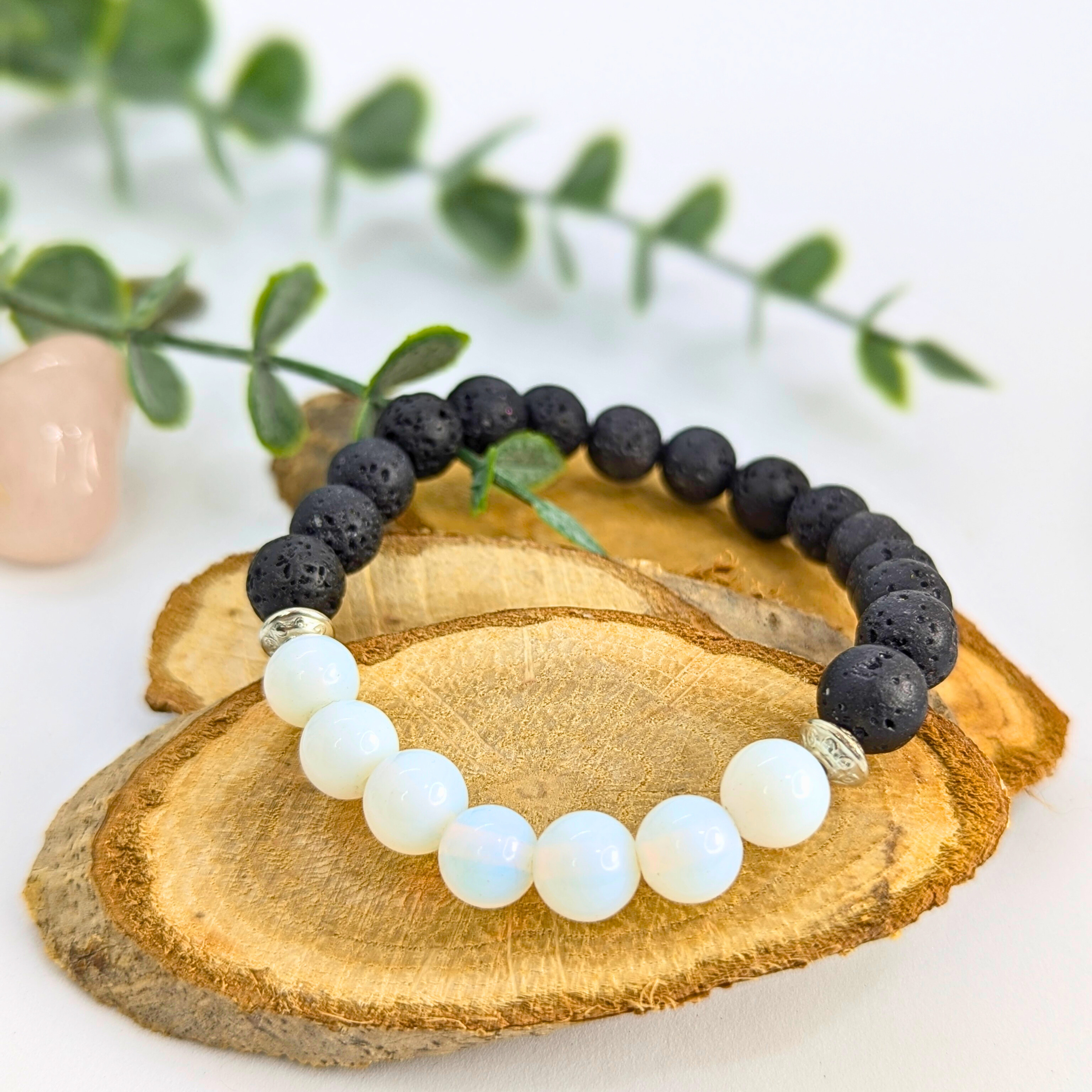 The Silver Sunbird Opal and Lava Stone Bracelet, featuring black lava stones and white opalite beads, is displayed on wooden slices with green leaves and a rose quartz crystal in the background.