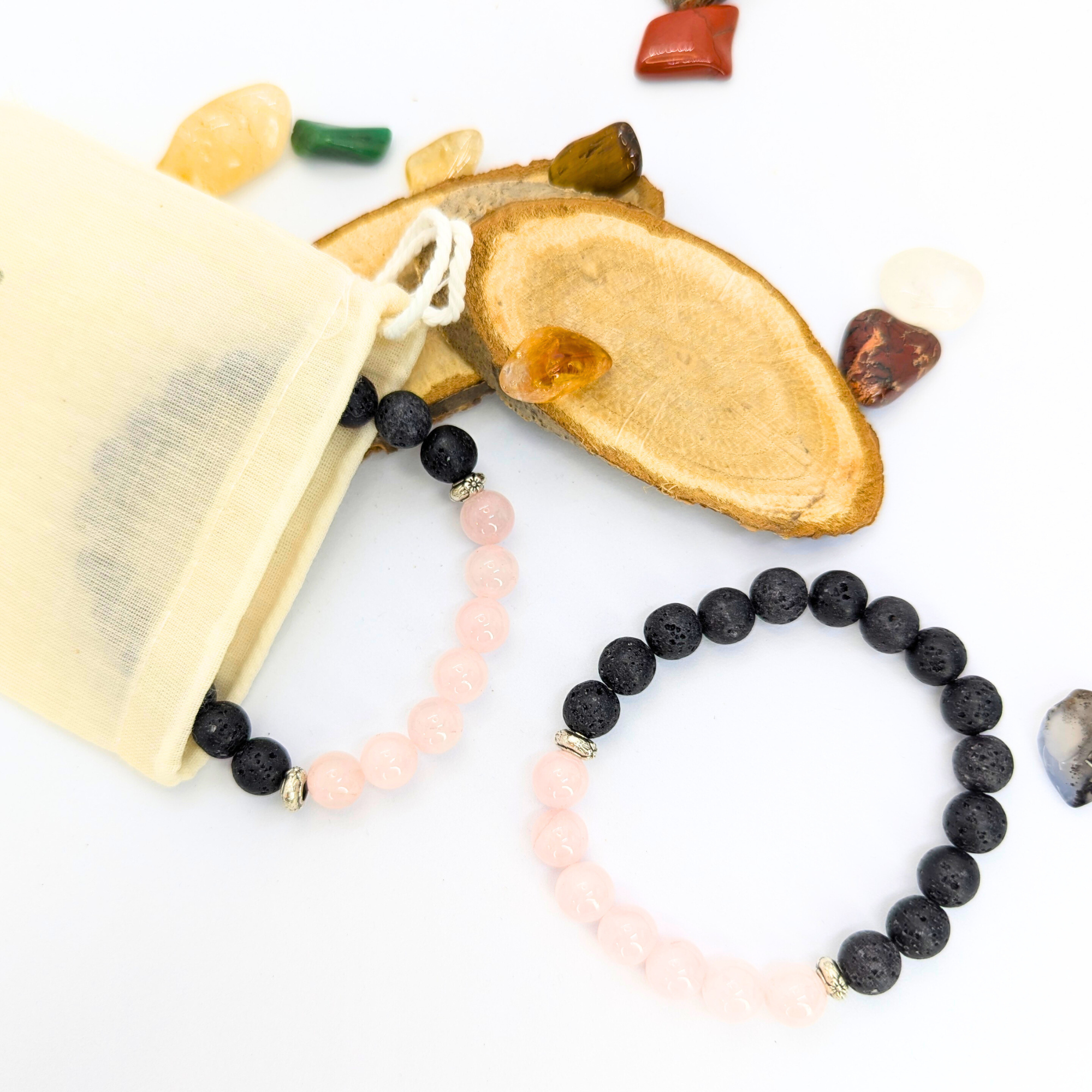 The Rose Quartz and Lava Stone Bracelets by Silver Sunbird are showcased on a white surface. Surrounding the bracelets are a cream-colored drawstring bag, wooden slices, and an array of scattered polished stones in various colors.