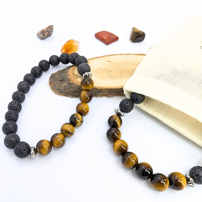 Two Silver Sunbird Tiger Eye and Lava Stone Bracelets are elegantly displayed on a light background beside a wooden slab and a beige pouch, with polished stones scattered nearby.