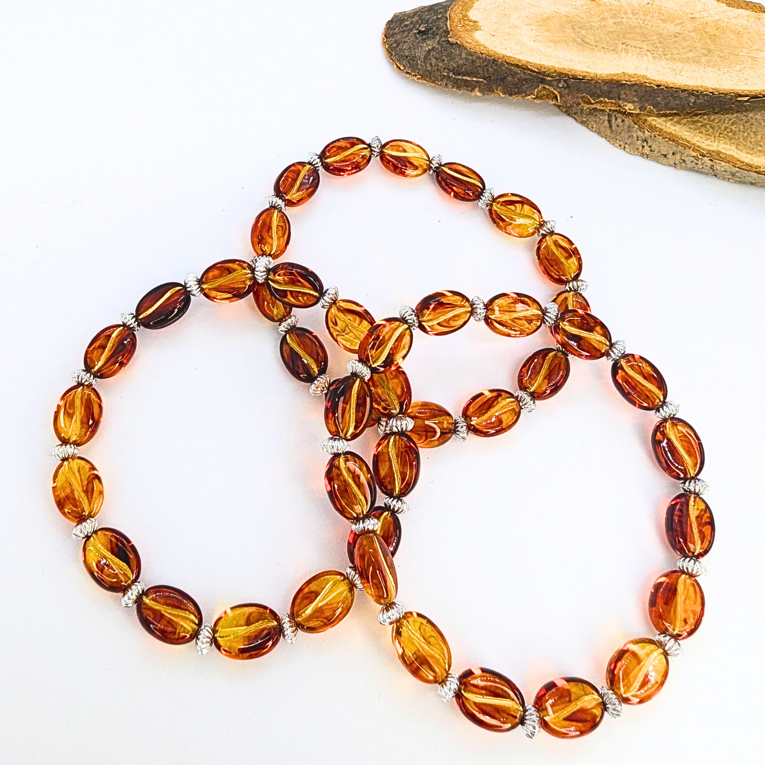 The Silver Sunbird Coffee Bean Bracelets, crafted from oval amber beads with silver spacers, are arranged in a circular pattern on a white surface. A wooden piece peeks out from the top right corner, adding an inviting touch for coffee lovers.