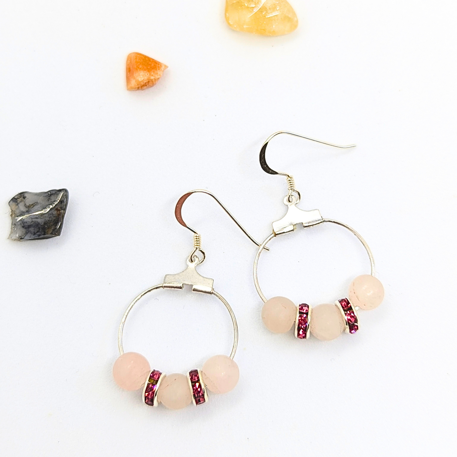 A pair of stunning Rose Quartz Statement Earrings by Silver Sunbird, featuring three round pink stones and red rhinestone spacers. These elegant hoop earrings, crafted from sterling silver, are set against a white background adorned with scattered colourful crystals in the corners, making them hypoallergenic for sensitive ears.