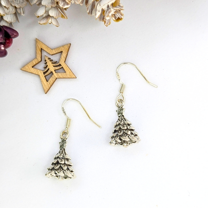 Two Silver Sunbird 3D Christmas Tree Earrings with hooks are displayed on a white surface next to a wooden star ornament with a cut-out tree design, plus dried flowers in the top left corner.