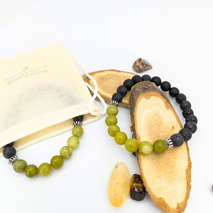 Two beaded bracelets from Silver Sunbird rest on wooden slices: one is a vibrant Peridot Bracelet with green beads, and the other has sleek black beads, like a lava stone bracelet. A small pouch and colorful stones are nearby, all set against a plain white background.