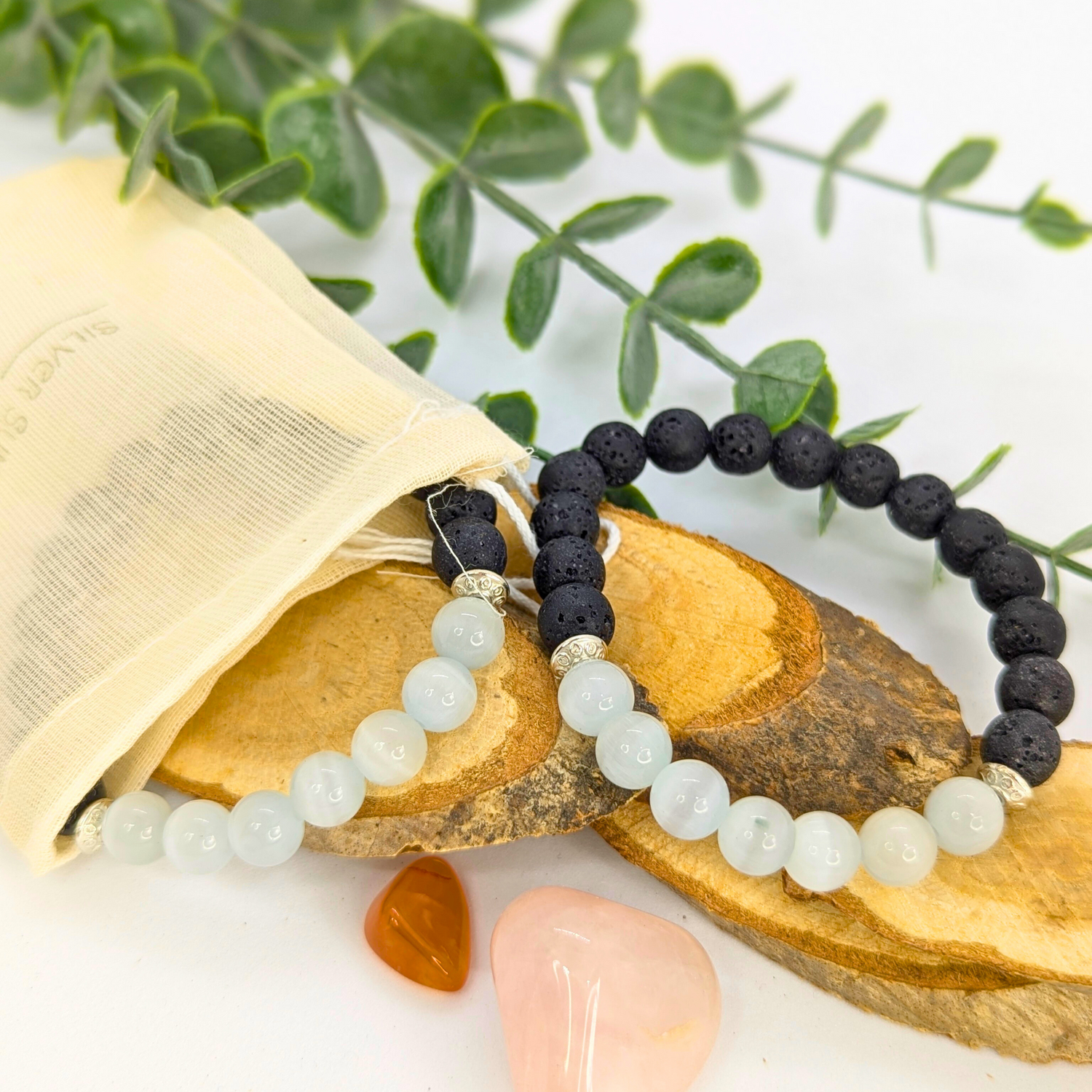 Silver Sunbird’s Moonstone and Lava Bracelets—one white moonstone, one black lava stone—are displayed on wooden slices with a cream pouch, greenery, and pink and orange stones arranged on a white background.