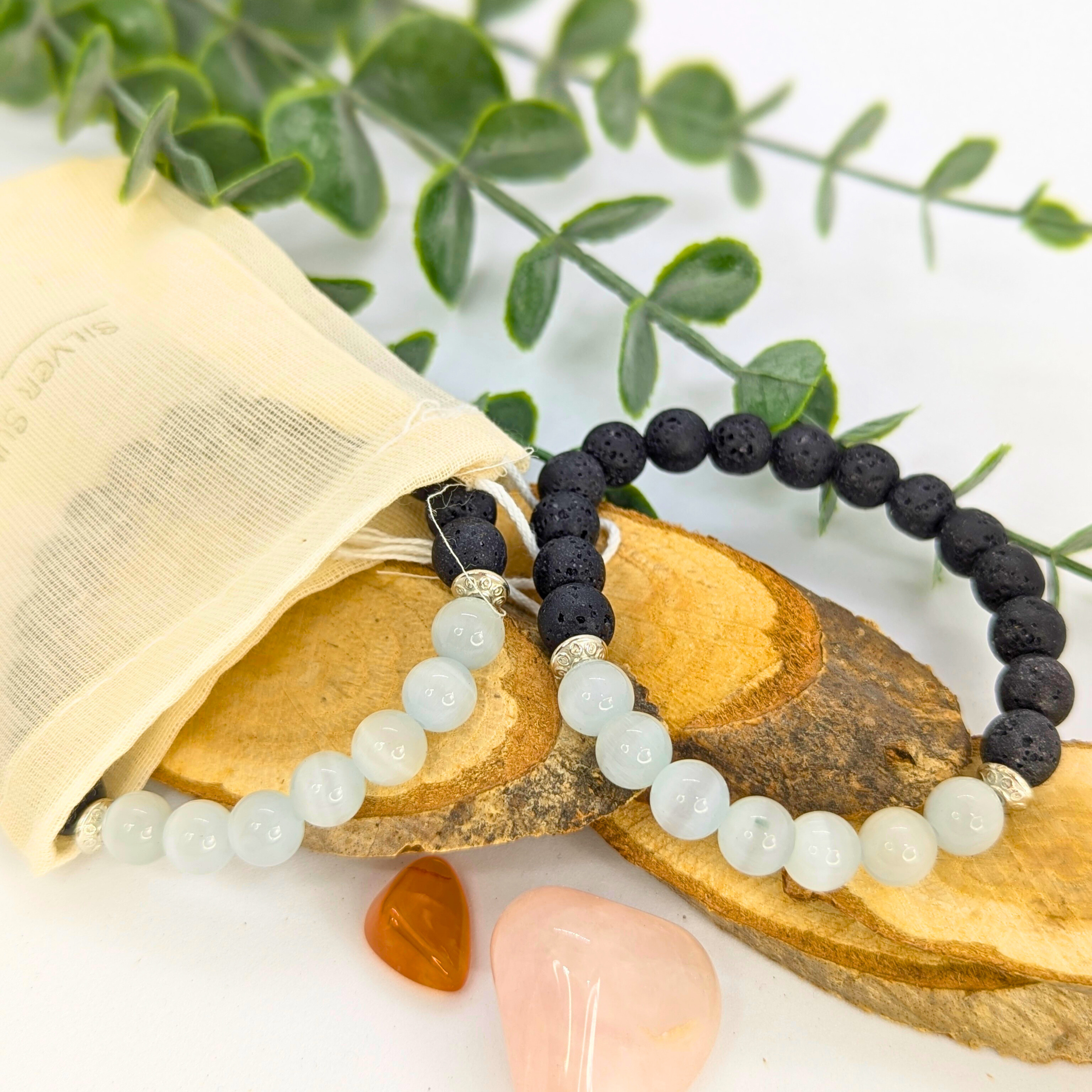 Silver Sunbird’s Moonstone and Lava Bracelets—one white moonstone, one black lava stone—are displayed on wooden slices with a cream pouch, greenery, and pink and orange stones arranged on a white background.
