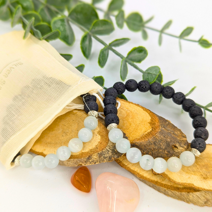 Silver Sunbird’s Moonstone and Lava Bracelets—one white moonstone, one black lava stone—are displayed on wooden slices with a cream pouch, greenery, and pink and orange stones arranged on a white background.