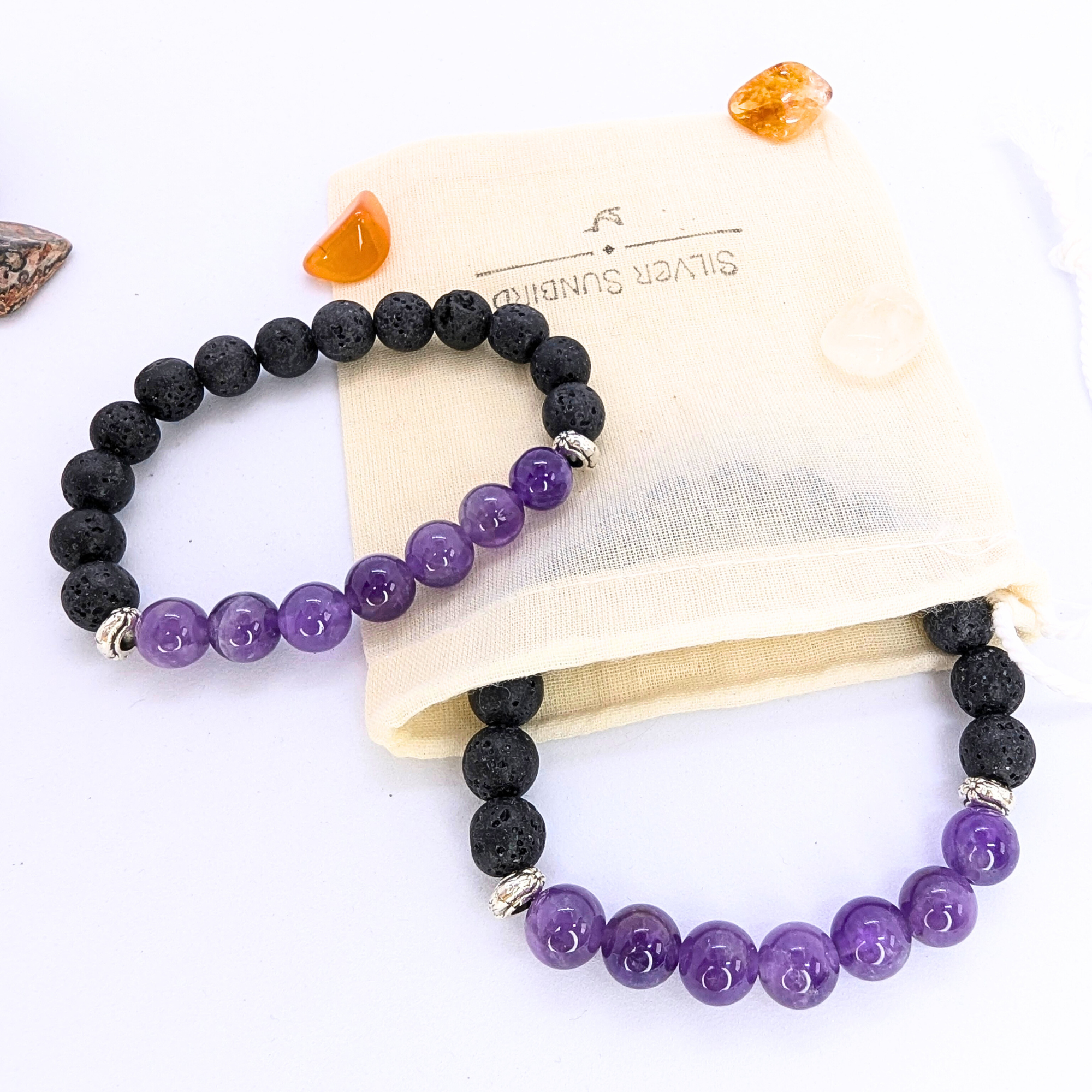 Two stretch bracelets featuring black lava stones and purple amethyst beads are displayed on a wooden slice. A beige drawstring pouch with &quot;Silver Surfer&quot; branding is positioned to the side, along with small polished stones in orange and brown hues. The amethyst beads are glossy and smooth, contrasting with the porous texture of the lava stones.