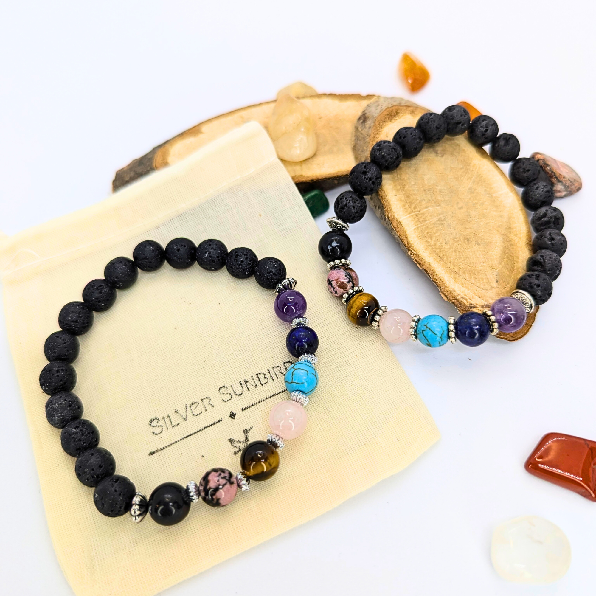 Two &quot;Silver Sunbird&quot; Chakra Bracelets, crafted with black and colorful semi-precious gemstones, rest on a cream pouch labeled with the brand name, surrounded by assorted stones and a piece of wood.