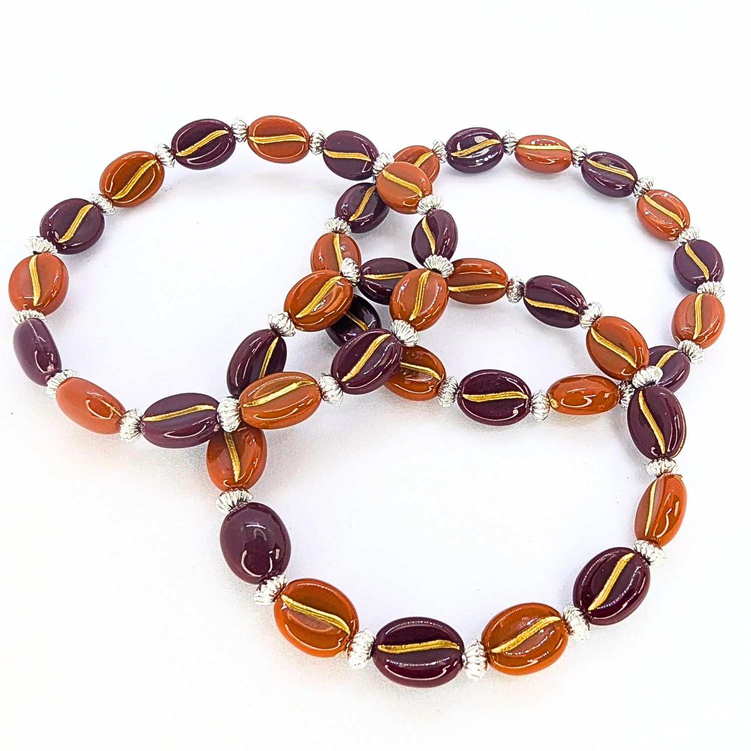 The Silver Sunbird Coffee Bean Bracelets feature three intertwined pieces with glossy orange and dark purple beads, detailed with gold stripes and separated by small silver beads, elegantly displayed on a plain white surface. Ideal for coffee lovers.