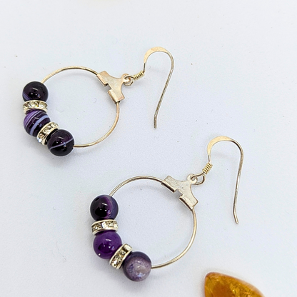 Introducing the Amethyst Statement Large Earrings by Silver Sunbird, an exquisite pair crafted from 925 sterling silver. These earrings feature a hook clasp and showcase alternating three amethyst gemstones and two silver rhinestone accents. A natural gemstone sits nearby on a white surface.