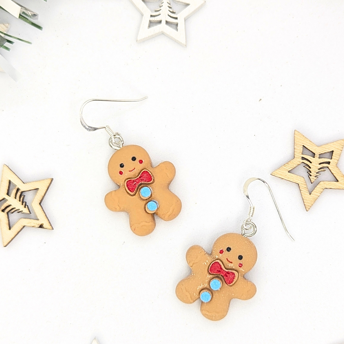 Two Cheerful Gingerbread Man Earrings by Silver Sunbird, featuring red and blue details, are arranged on a white surface, surrounded by wooden star ornaments.