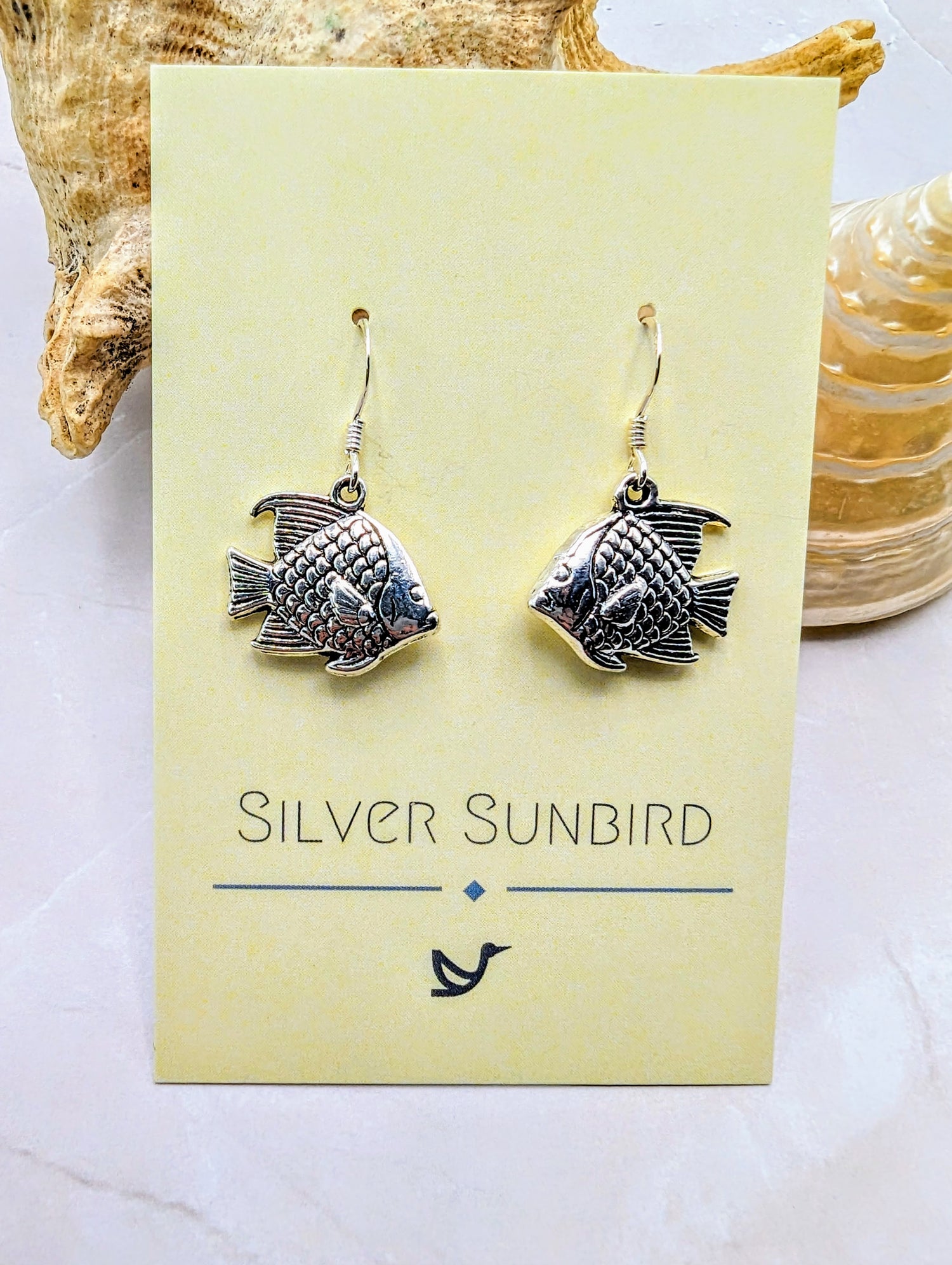 A pair of Angelic Angelfish Earrings, crafted from sterling silver, is displayed on a yellow card with the brand name &quot;Silver Sunbird.&quot; The dangle earrings, perfect for any ocean lover, is set against a backdrop that includes seashells and a light-coloured surface.