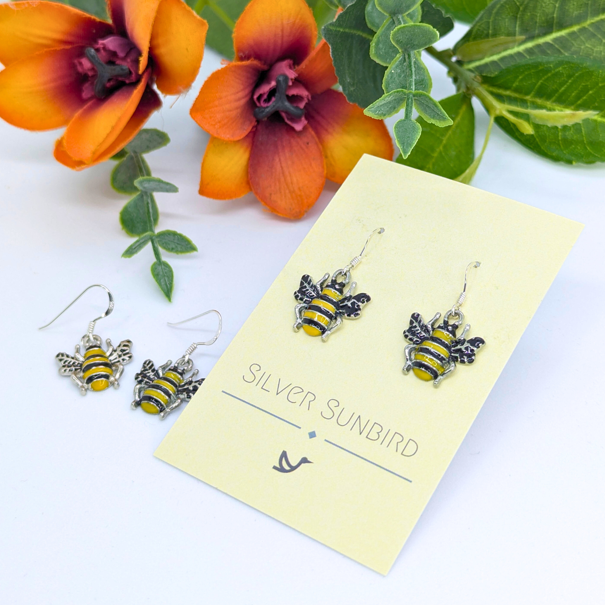Bumble Bee Earrings by Silver Sunbird, featuring yellow and black stripes, are shown on a branded card alongside another pair with orange flowers and green leaves in the background.