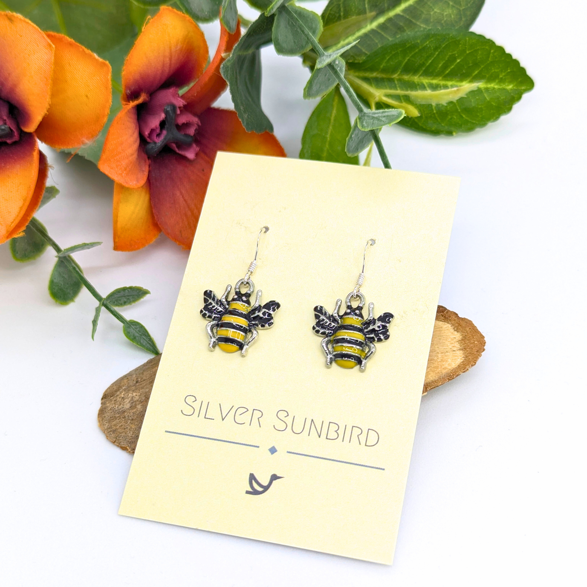 Bumble Bee Earrings by Silver Sunbird, featuring yellow and black enamel stripes, are displayed on a branded card atop wood, accented by orange flowers and green leaves for a charming presentation.