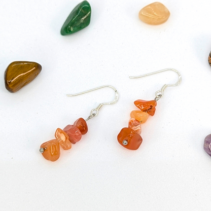 Silver Sunbird Carnelian Agate Earrings feature orange gemstone chips on sterling silver dangles, displayed on white and surrounded by colorful polished stones.