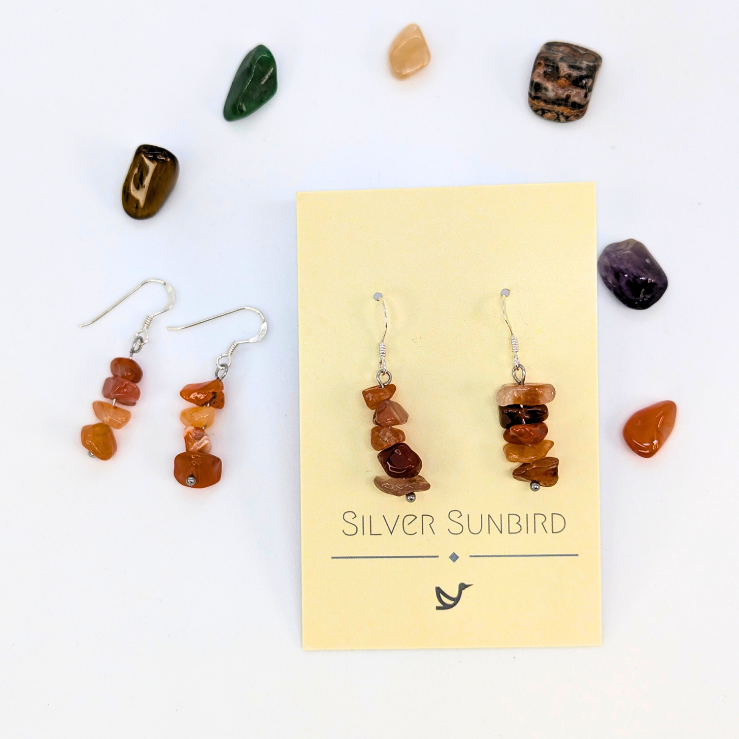 Carnelian Agate Earrings by Silver Sunbird: handmade sterling silver dangle earrings with stacked brown stones, displayed on a branded card and surrounded by polished gemstones—a unique accessory for any occasion.