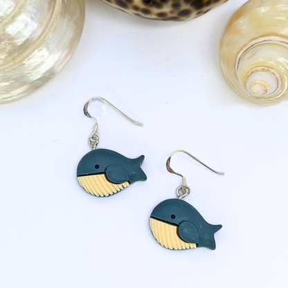 Colourful Whale Earrings by Silver Sunbird, featuring dark blue bodies and beige bellies, are displayed on a white surface with two light-colored seashells.
