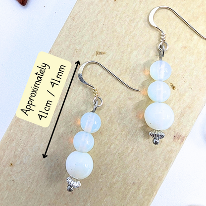 The Opal Dangle Earrings by Silver Sunbird have graduated white beads on silver hooks, measuring about 4.1 cm or 41 mm, and are displayed on a wooden surface.